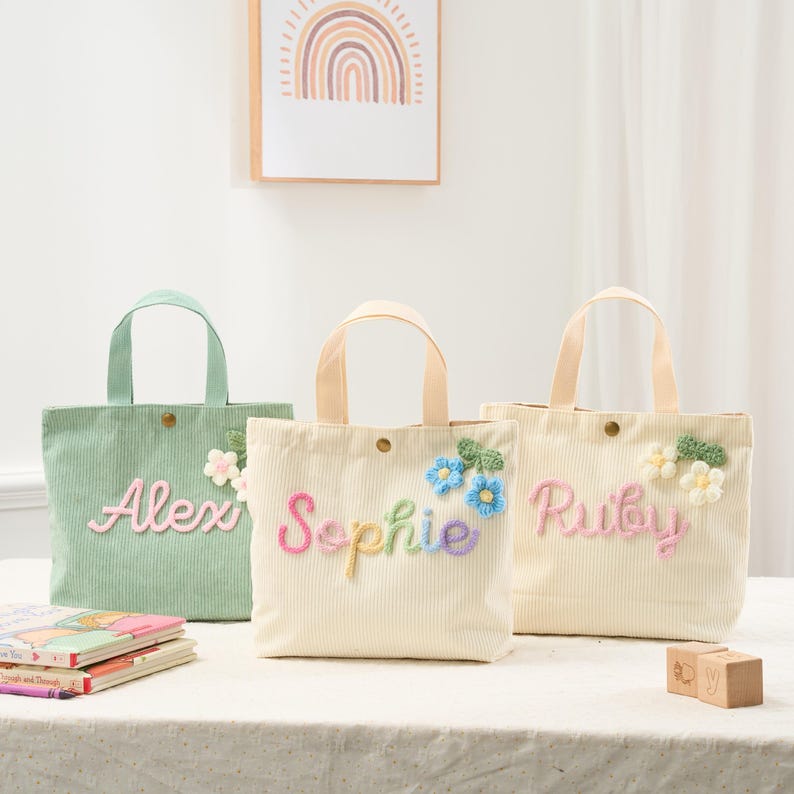 May include: Three corduroy tote bags in pastel colors, each with a name embroidered in colorful thread. The bags have matching handles and floral accents. The names are Alex, Sophie, and Ruby.