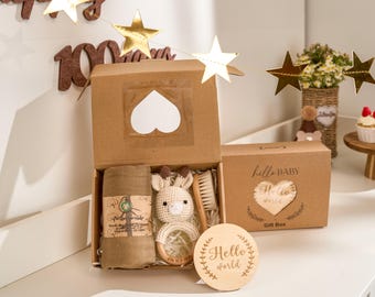 May include: A baby gift box with a heart-shaped window, containing a muslin swaddle, a crochet giraffe rattle, a wooden brush, and a wooden teething ring. The box also includes a wooden sign that says "Hello world".