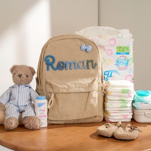 May include: A tan corduroy backpack with the name "Roman" in blue letters, a teddy bear, baby powder, diapers, wipes, and baby shoes are arranged on a wooden surface. The backpack has a small blue bow.