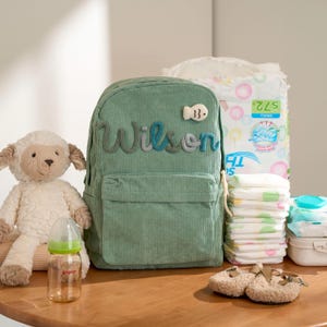 May include: A sage green corduroy backpack with the name "Wilson" embroidered on the front, next to a fish-shaped patch. A plush lamb, baby bottle, diapers, wipes, and baby shoes are also visible.