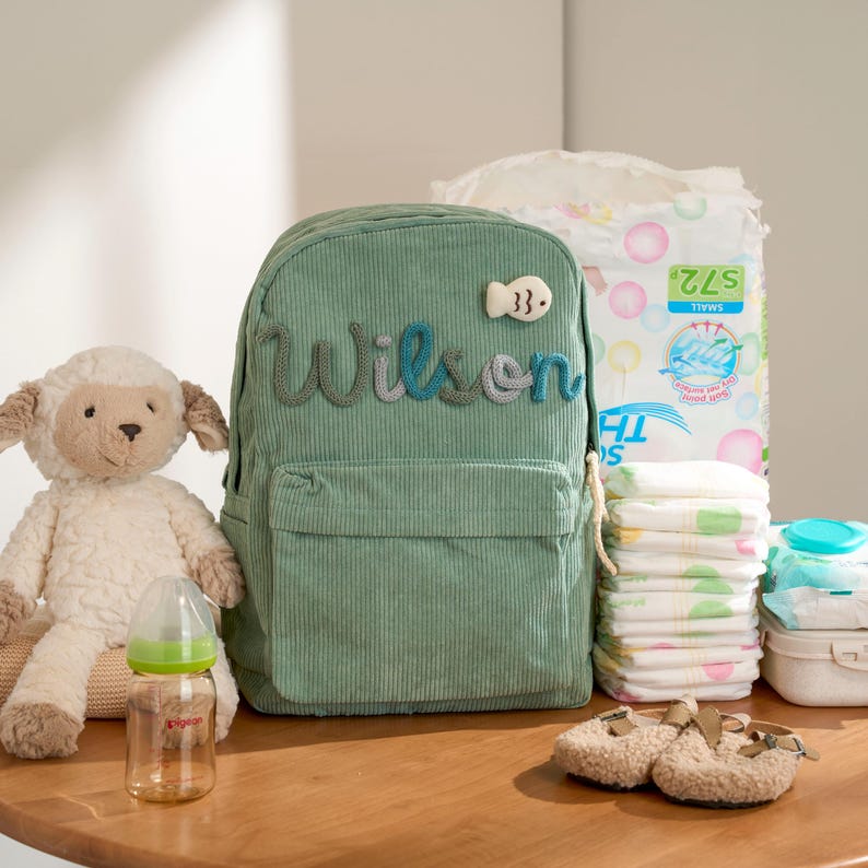 May include: A sage green corduroy backpack with the name "Wilson" embroidered on the front, next to a fish-shaped patch. A plush lamb, baby bottle, diapers, wipes, and baby shoes are also visible.