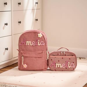 Peut inclure: Un sac à dos en velours côtelé rose et un sac à déjeuner assorti. Le sac à dos a une poche avant et un motif floral. Les deux articles sont personnalisés avec le nom "Amelia".