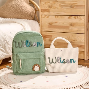 May include: A green corduroy backpack and a white tote bag. Both bags have the name "Wilson" embroidered on them in a mix of colors. The backpack features a small hedgehog patch. The bags are on a round rug.