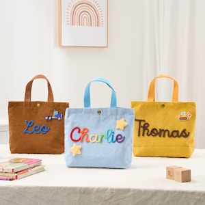 May include: Three corduroy tote bags in brown, light blue, and yellow. Each bag has a name embroidered on it: "Leo", "Charlie", and "Thomas". The "Leo" bag has a truck patch, the "Charlie" bag has star patches, and the "Thomas" bag has a construction vehicle patch.