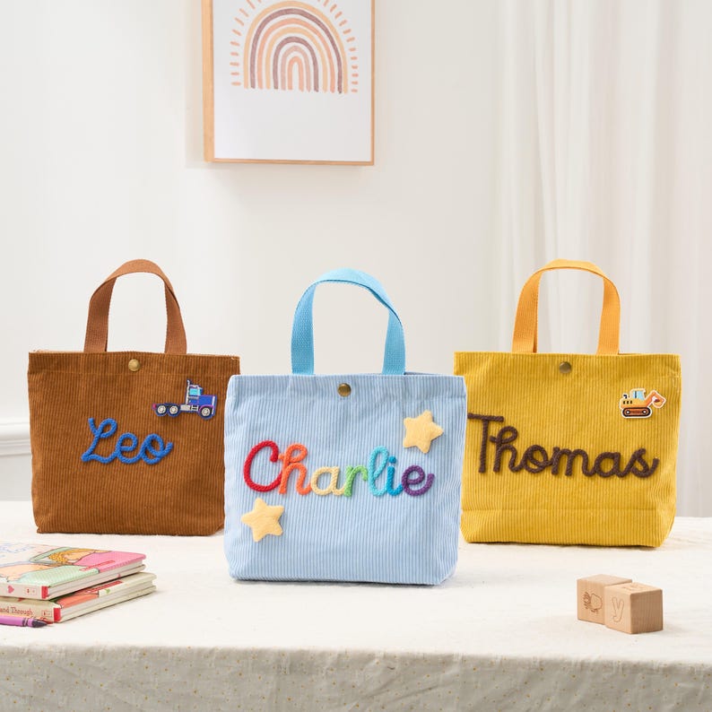 May include: Three corduroy tote bags in brown, light blue, and yellow. Each bag has a name embroidered on it: "Leo", "Charlie", and "Thomas". The "Leo" bag has a truck patch, the "Charlie" bag has star patches, and the "Thomas" bag has a construction vehicle patch.