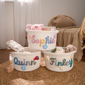Op de afbeelding: Drie witte touwmanden met geborduurde namen en bloemaccenten. De manden zijn gevuld met speelgoed en dekens. De namen "Sophie", "Quinn" en "Finley" zijn geborduurd met kleurrijk garen. Op één mand zit een rode auto-applicatie.