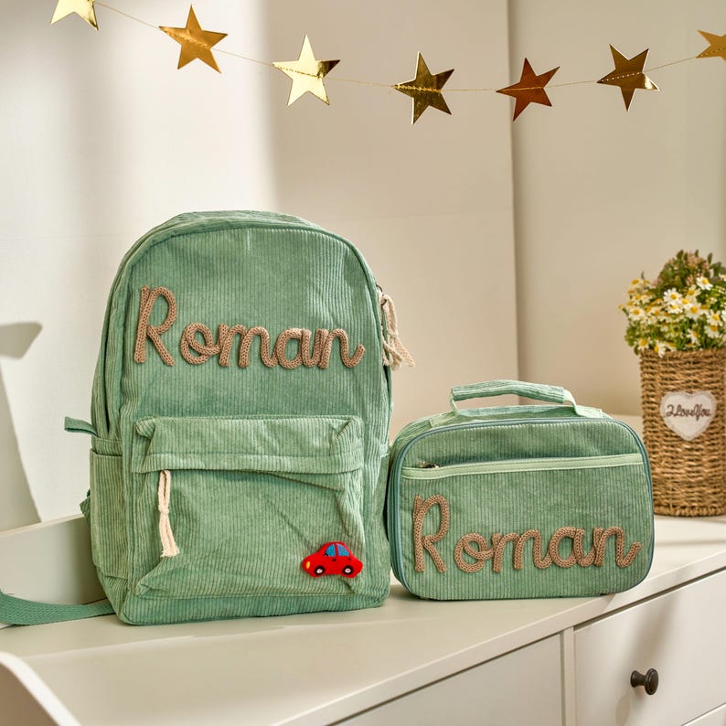 Op de afbeelding: Een groene corduroy rugzak en bijpassende lunchtas. De rugzak en lunchtas hebben de naam "Roman" geborduurd in een bruin script. Een kleine rode auto-applicatie staat op de rugzak. Gouden sterrenslingers op de achtergrond.