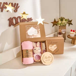 Könnte beinhalten: Ein Baby-Geschenkset in einem Karton, einschließlich einer rosa Wickeldecke, einem Häschen-Beißring, einer Bürste und einem Holzschild mit der Aufschrift "Hello World". Die Box ist mit "Hello Baby" beschriftet. Goldene Sterne als Dekoration im Hintergrund.