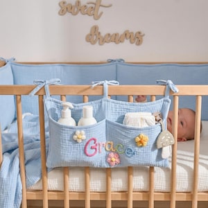 May include: A light blue baby crib organiser with three pockets, embroidered with the name "Grace" in colourful thread and decorated with flower appliqués. The organiser holds baby essentials and is attached to a wooden cot.
