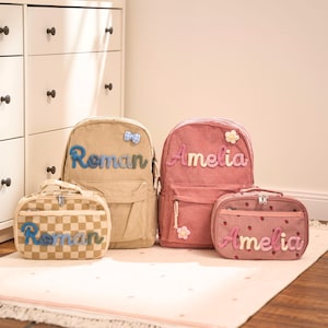 May include: Four corduroy bags, including two backpacks and two smaller cases. One backpack is beige with the name "Roman" in blue, and the other is pink with "Amelia" in pink. The smaller bags match the colours and names.