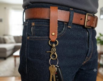 May include: A tan leather belt loop keychain holder featuring a brass-toned swivel lobster clasp and a circular split ring. The leather strap is secured with a brass rivet and hangs from a brown leather belt worn over dark blue denim jeans.