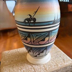 Peut inclure: Un vase en céramique peint à la main avec un motif amérindien. Le vase présente une silhouette d'un cavalier à cheval sur fond de coucher de soleil. Le vase a une base blanche et est décoré de motifs géométriques et de bandes de couleurs.