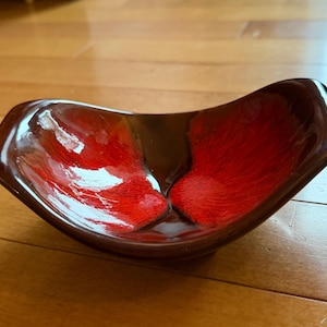 May include: A small, curved, boat-shaped bowl with a glossy finish. The interior features a vibrant, textured red pattern that fades into a deep, dark brown border. The exterior and edges are a solid, dark, reflective brown color.