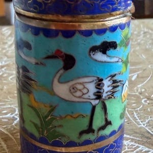 Puede incluir: Un recipiente cilíndrico de cloisonné con tapa extraíble, que presenta un fondo de color azul claro y verde vibrante decorado con grullas blancas, detalles en negro y filigrana de metal dorado. Los bordes superior e inferior son azul oscuro.