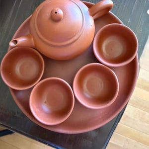 Puede incluir: Un juego de té de terracota que consta de una tetera redonda con tapa y cuatro cuencos pequeños a juego, todo dispuesto sobre una bandeja circular de terracota. El conjunto presenta un acabado mate de color marrón rojizo uniforme y formas redondeadas.
