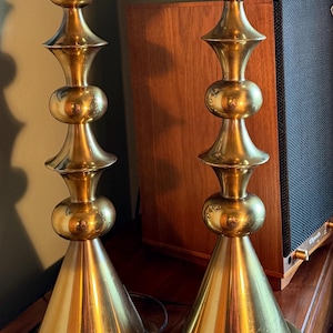 May include: Two vintage brass table lamps. Each lamp has a conical base, a series of rounded and hourglass-shaped sections, and a cylindrical top. The lamps are gold-coloured and reflect light, suggesting a polished finish. The lamps are placed on a wooden surface.