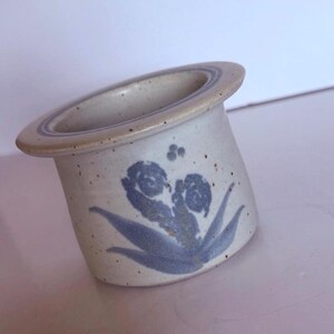 May include: A speckled off-white ceramic egg cup with a wide, flat rim featuring a subtle blue circular band. The exterior body is decorated with a blue painted floral motif consisting of two curled fern-like shapes and small dots above.