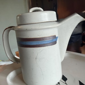 May include: A cylindrical ceramic teapot with a speckled off-white glaze, featuring a horizontal band of dark brown and blue stripes. The teapot includes a matching lid with a small handle, a curved side handle, and a wide, angular pouring spout.