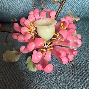 Könnte beinhalten: Eine rosa-grüne Blumenskulptur mit einem zentralen, hellgrünen, becherförmigen Halter. Die Skulptur zeigt rosa Blütenblätter, gelbe Akzente und grüne Blätter. Die Skulptur besteht aus einem durchscheinenden Material und hat einen Metallstiel.