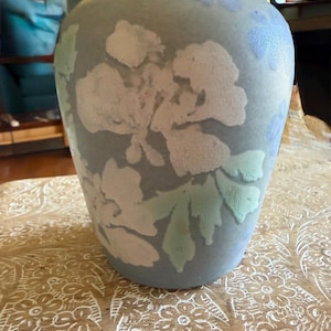 May include: A matte, light blue ceramic vase featuring a textured, raised floral pattern in shades of white, pale green, and soft lavender. The vessel has a rounded, tapered silhouette with a wide opening and a smooth, consistent finish throughout.