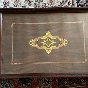 May include: A rectangular wooden serving tray featuring a dark brown finish with a central gold-colored decorative emblem. The tray includes two side handles with metallic accents and a thin, light-colored border framing the central design.