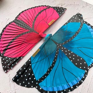 May include: Two folding hand fans shaped like butterfly wings. One fan is vibrant pink with black edges, white dots, and black vein lines. The other fan is bright blue with black edges, white dots, and black vein lines. Both feature gold metal accents.