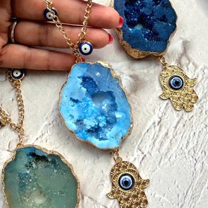 May include: Three wall hangings featuring gold-tone chains adorned with small blue and white evil eye beads. Each chain holds a large, irregular geode slice in shades of blue or green, with a gold-tone hamsa hand charm featuring an evil eye bead.