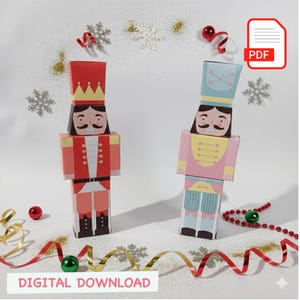Puede incluir: Dos figuras de cascanueces de papel, una roja y otra rosa, sobre una superficie blanca con adornos navideños. El cascanueces rojo lleva corona y uniforme. El rosa tiene sombrero azul y uniforme rosa. Las palabras "DIGITAL DOWNLOAD" están en la parte inferior.
