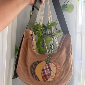 May include: A tan corduroy hobo bag featuring a quilted diamond pattern and an olive green adjustable strap. The front is decorated with a large, multi-patterned fabric pumpkin appliqué in shades of brown, yellow, and orange plaid and floral prints.