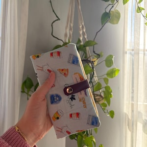 May include: A light gray fabric pouch featuring a repeating pattern of coffee cups, pizza slices, and takeout boxes. The pouch is secured with a dark purple leather strap and a metallic snap button. A small fabric tag with text is visible on the front.