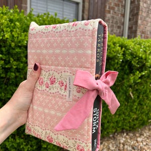 May include: A pink and cream floral fabric book cover with a matching bow closure. The cover features a quilted design with a floral and lace pattern. The book title "Onyx Storm" is visible.