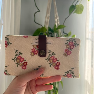 May include: A rectangular quilted fabric wallet featuring a cream background with a repeating pattern of small pink and yellow flowers with green leaves. A dark brown leather strap with a metal snap closure secures the center, featuring a small white fabric label with the text "cro...aisies".