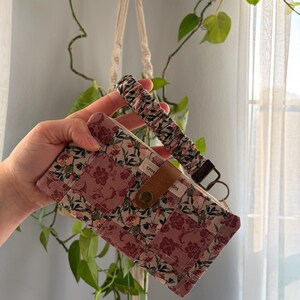 May include: A rectangular fabric wallet featuring a patchwork design of floral patterns in shades of dusty rose, cream, and sage green. It includes a brown leather snap closure tab and a matching gathered fabric wrist strap with a metal key ring.