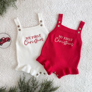 May include: Two knitted baby rompers, one white and one red, with the text "MY FIRST Christmas" embroidered in red. Each romper has button fastenings and frilled edges. A Christmas-themed coaster is also visible.