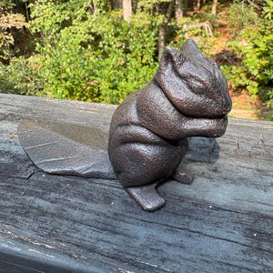 May include: A dark brown, textured cast iron squirrel figurine depicted in a seated position with its front paws held near its face. The squirrel features a prominent, flat, leaf-shaped tail extending horizontally behind its body.