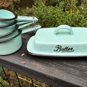 May include: A set of light mint green enamel measuring cups with black rims, stacked and held together by a metal ring, alongside a matching rectangular butter dish with a lid featuring the word Butter in black script lettering.