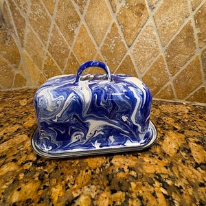 May include: A rectangular butter dish with a lid featuring a blue and white marbled pattern. The lid has a curved handle on top, and the entire piece sits on a matching rectangular base with a silver-toned rim, showcasing a glossy finish.
