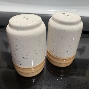 May include: A pair of cylindrical salt and pepper shakers featuring a white speckled glaze on the upper portion and a smooth, unglazed tan base. Each shaker has a flat top with three small holes for dispensing seasoning in a vertical orientation.