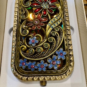 May include: A rectangular gold-toned metal compact mirror featuring an ornate floral overlay. The design includes red and blue enameled flowers with embedded crystals, set against a textured gold frame with swirling filigree accents.
