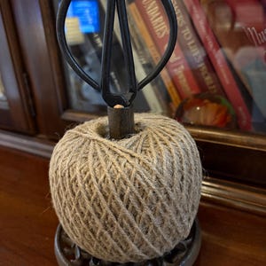 May include: A twine dispenser with a ball of natural-colored twine. The twine ball sits on a decorative, dark metal base. A pair of black scissors is mounted on top, designed for cutting the twine. The background includes a bookshelf with books.
