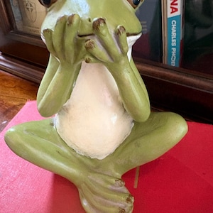 May include: A green frog figurine sitting in a cross-legged position with its hands covering its mouth. The frog has a cream-colored belly, dark eyes, and textured skin. The figure is depicted in a pose resembling the speak-no-evil gesture.