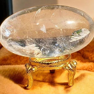 May include: A clear quartz crystal sphere, displayed on a decorative gold-coloured stand. The sphere has internal fractures and inclusions, and the stand features ornate detailing. The overall aesthetic is elegant and decorative.