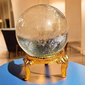 May include: A clear crystal ball with a textured surface, on a gold-coloured, ornate stand with four legs. The stand is placed on a blue surface. The crystal ball is a decorative item.