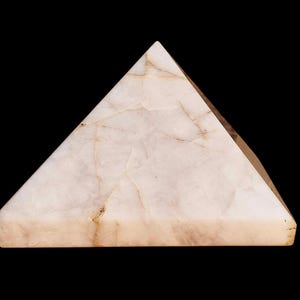 May include: A pyramid-shaped ornament crafted from a pale, translucent stone. The stone features a creamy white hue with delicate brown veining, and a polished finish. The pyramid is displayed against a black backdrop.