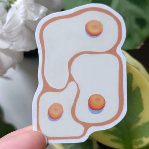 May include: A die-cut sticker featuring a design of three fried eggs. The eggs have white egg whites with orange yolks and are outlined in a light brown colour. The sticker is on a white background.