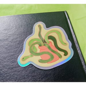May include: A holographic sticker showing a cartoon illustration of green snakes coming out of a pink pouch. The sticker has a yellow background and a shiny, iridescent border. The sticker is placed on a dark surface.