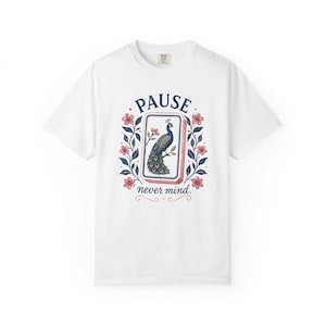 Könnte beinhalten: Ein weißes Kurzarm-T-Shirt mit einer zentralen Grafik eines Mahjong-Steins mit Pfauenmotiv. Der Text PAUSE steht über dem Stein und never mind. darunter, umrahmt von einem dekorativen Kranz aus rosa Blüten und blauen Blättern auf dem Stoff.