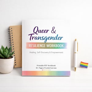 Peut inclure: Un cahier blanc intitulé "Queer & Transgender Resilience Workbook" avec une bande arc-en-ciel en bas. Un stylo, un petit drapeau arc-en-ciel, une plante succulente et un carnet à spirales sont à côté du livre.