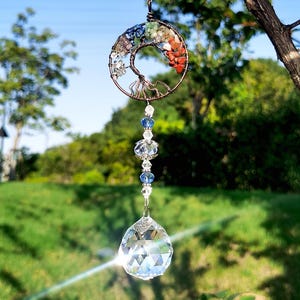May include: A decorative suncatcher featuring a wire tree of life design, adorned with colorful gemstones and a large, faceted crystal ball. The suncatcher hangs from a thin string, with blue and clear beads. The background is a blurred view of green trees and a blue sky.