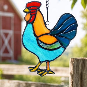 Peut inclure: Un attrape-soleil en vitrail en forme de coq, composé de panneaux de verre aux couleurs vives bleu, orange, rouge et jaune, assemblés par des lignes de soudure en métal noir. Le coq est suspendu à une chaîne en métal noir, offrant un design translucide.